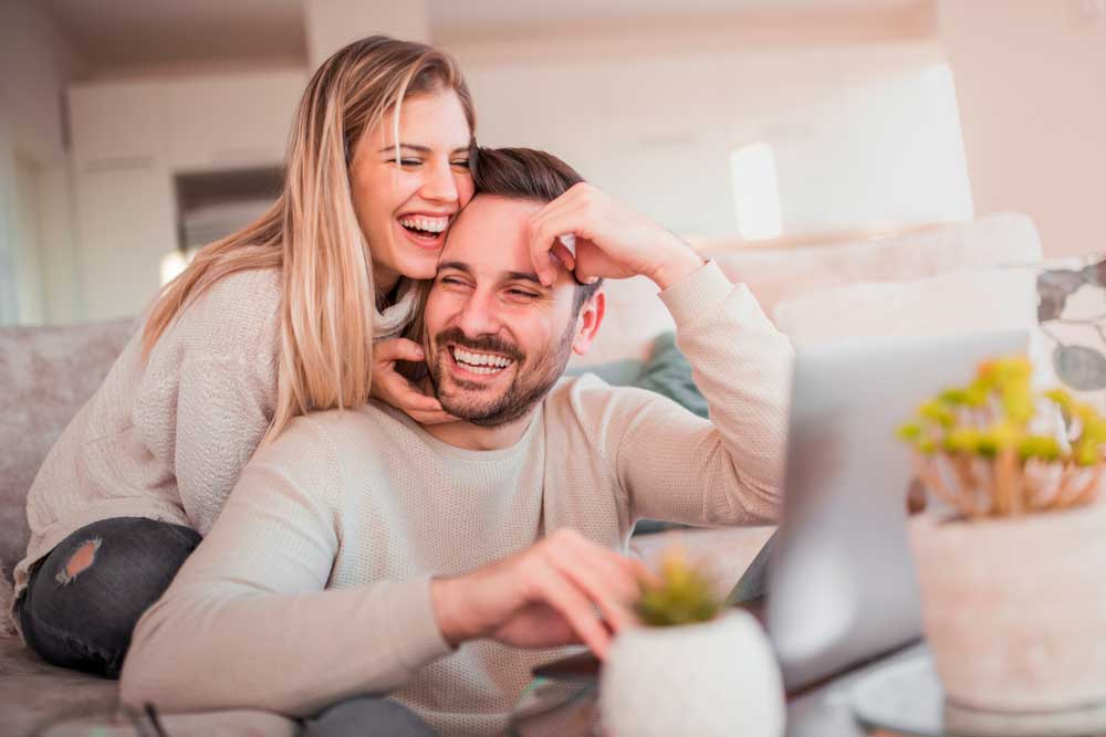 happy couple with a girl hugging a guy, holding his face and both smiling, guy using a laptop