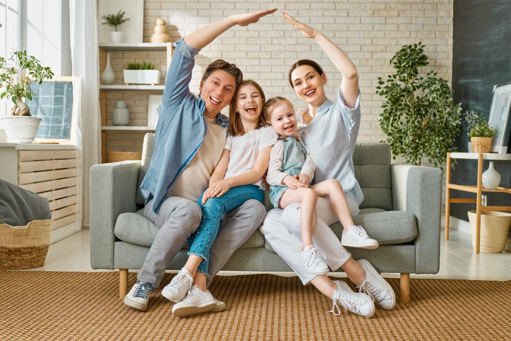 Happy family sitting on a grey couch with their two kids making a house roof with their hands
