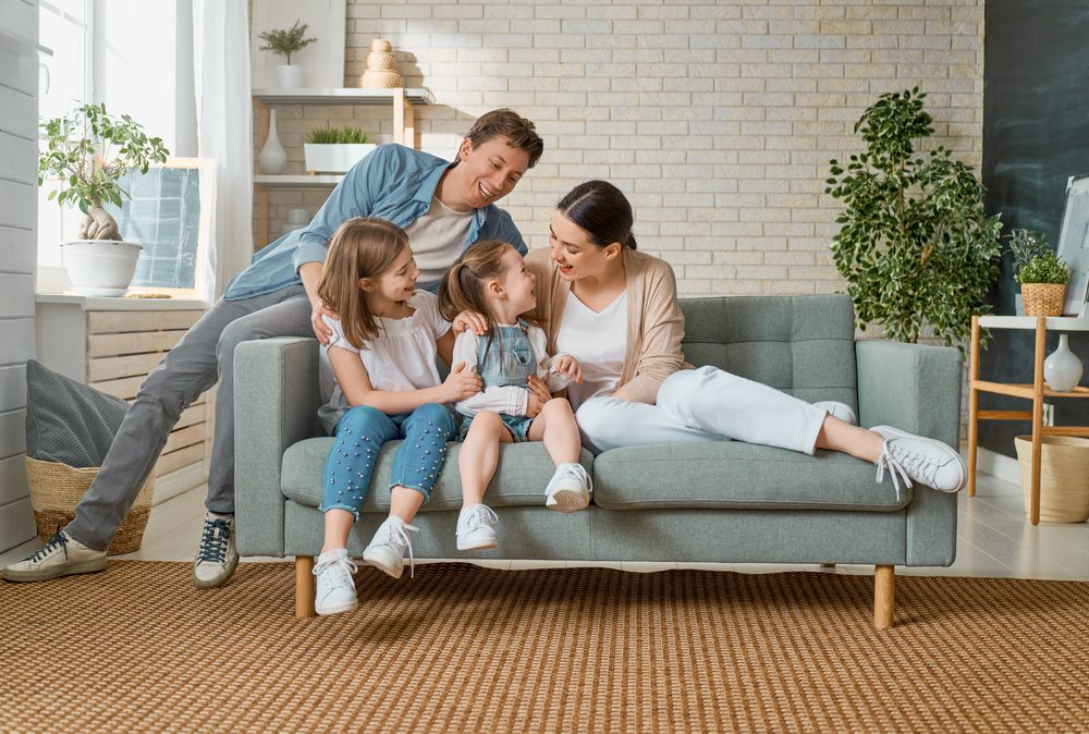 A happy family with a husband and wife and two kids sitting on a grey couch with a huge tree in the back and lots of plants
